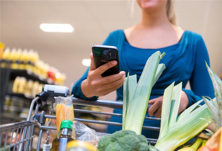 Eine Frau hält ein Smartphone neben ihrem Einkaufswagen in einem Supermarkt in der Hand.