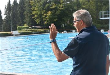 Mann mit Brille steht am Schwimmbecken und winkt.