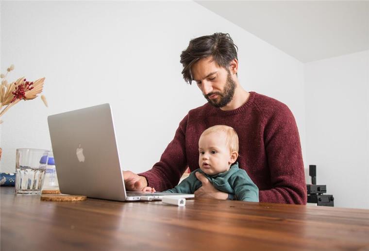 Ein Mann, mit einem Baby auf dem Schoss, sitzt an seinem Schreibtisch und macht Homeoffice. 