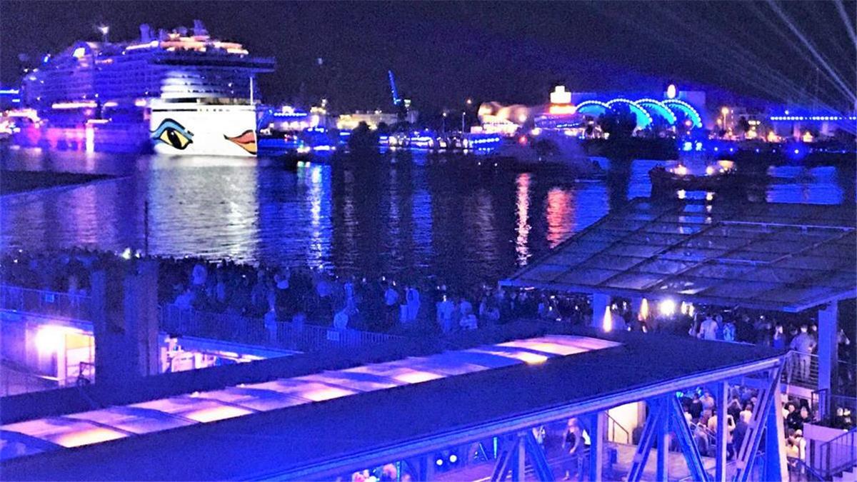 Hamburg bleibt Kreuzfahrt-Hotspot: Aida und Co. sorgen für Rekordzahlen Nachtaufnahme eines Hafens mit einem großen, beleuchteten Kreuzfahrtschiff im Hintergrund, das das charakteristische Design von Augen und Lippen auf der Schiffswand trägt. Der Hafen ist in blauem Licht illuminiert, mit zahlreichen Menschen, die das Szenario von einer Aussichtsplattform und einer unteren Ebene beobachten.