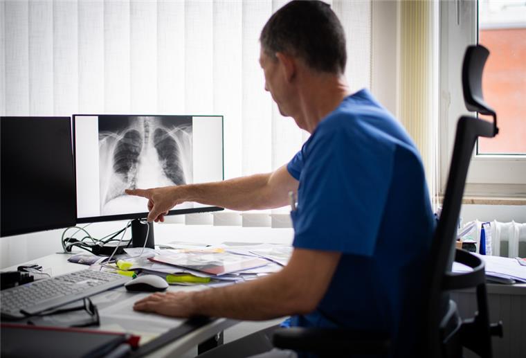 Ein Arzt im blauen Kasack zeigt auf einen Monitor. Zu sehen ist eine Röntgenaufnahme einer entzündeten Lunge.