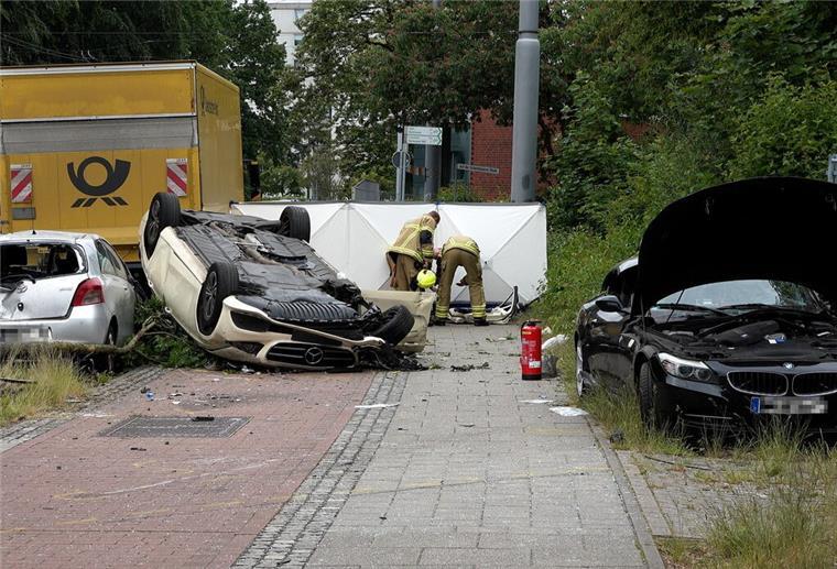 Ein stark beschädigter Taxi-Wagen liegt auf dem Dach nahe einer Straße. Weitere Fahrzeuge stehen mit deutlichen Schäden in der Nähe.
