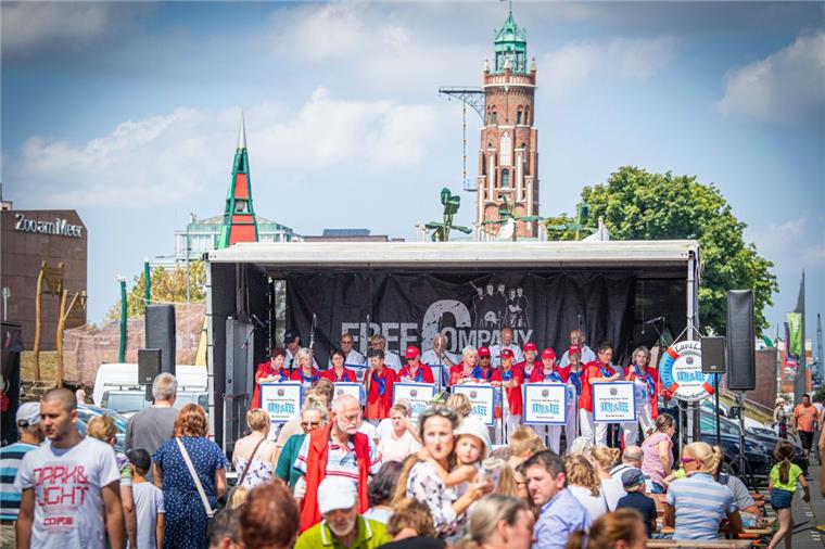 Besucher bummeln über das Leuchtturmfest am Neuen Hafen in Bremerhaven und lauschen  dem Chor "Luv & Lee" auf der Bühne. 