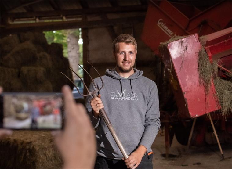 Landwirt aus dem Cuxland ist ein YouTube-Star Ein Mann steht mit Forke in einer Scheune, im Vordergrund ist unscharf ein Handydisplay zu sehen.
