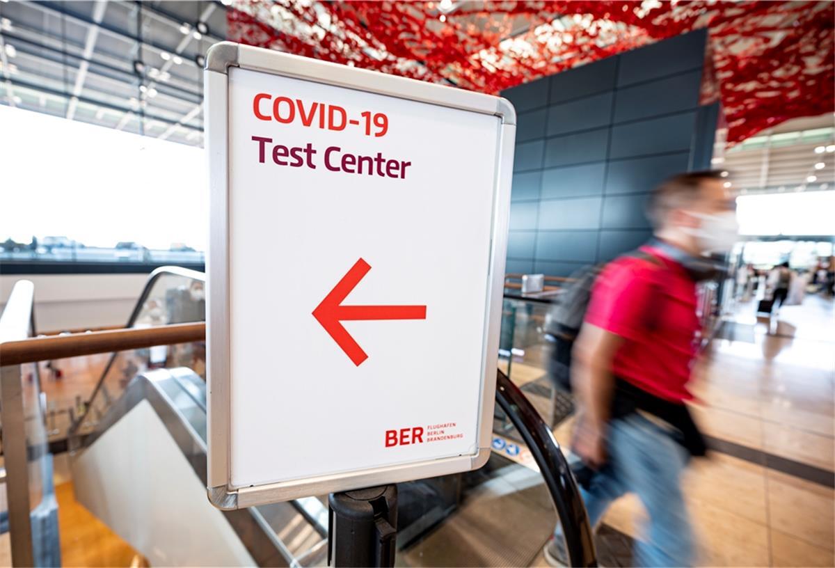 Weil und Kretschmann für kostenpflichtige Corona-Tests Fluggäste laufen durch die Haupthalle des Flughafens Berlin Brandenburg (BER) an einem Schild mit der Aufschrift "Covid-19 Test Center" vorbei.