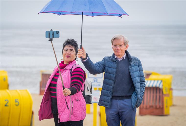 YouTuber aus Cuxhaven begeistern weltweit mit Urlaubsvideos Eine Frau und ein Mann stehen am Strand von Duhnen. Sie hält ein Smartphone auf einem Selfiestick, er den Regenschirm.