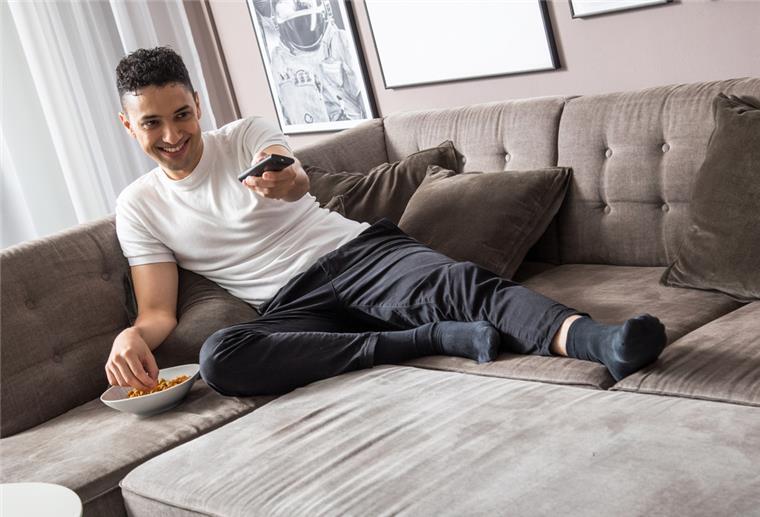 Gemütlich auf der Couch: Streaming-Tipps für‘s Wochenende Ein Mann sitzt mit Snacks und einer Fernbedinung in der Hand auf der Couch.