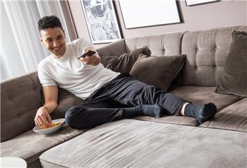 Ein Mann sitzt mit Snacks und einer Fernbedinung in der Hand auf der Couch.