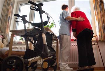 Eine Pflegerin im Heim hilft einer Seniorin, die stehen aus einem Fenster guckt.
