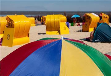 Ein bunter Sonnenschirm im Vordergrund, dahinter Sandstrand, gelbe Strandkörbe und Menschen, verschwommen.