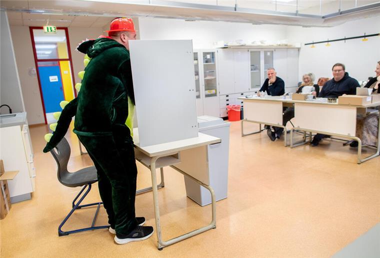 Dinosaurier, Elvis und Co. stürmen das Wahllokal! Der kostümierte Wähler Christopher steht in einem Wahllokal bei der Stimmabgabe hinter einem Sichtschutz.