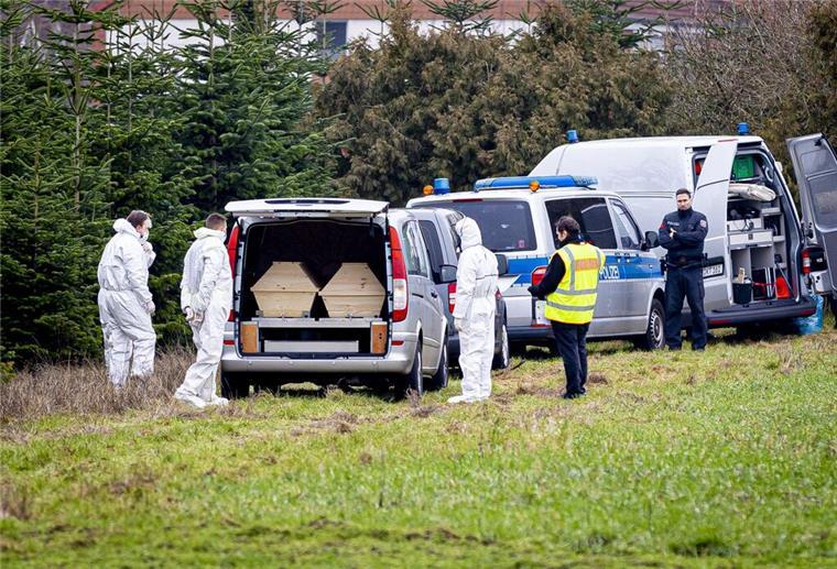 Mitarbeiter der Spurensicherung der Polizei stehen an einem Leichenwagen, der auf einer Wiese parkt. 