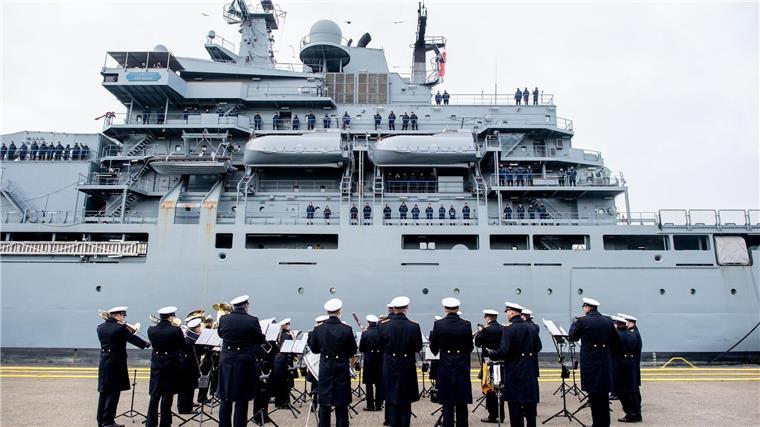 Der Marinemusikkorps Wilhelmshaven steht an einer Kaimauer, während die „Frankfurt am Main“ in den Hafen einfährt.