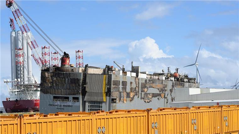 Fremantle Highway: Ladung wird nach Feuer vom Autofrachtschiff geborgen Der schwer beschädigte Autofrachter “Fremantle Highway„ liegt im Hafen, abgeschirmt durch gelbe Container.