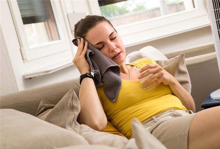 Cool bleiben im Dachgeschoss: Tipps gegen die Sommerhitze Eine Frau auf dem Sofa mit einem Handtuch am Kopf und einem Glas Wasser in der Hand.