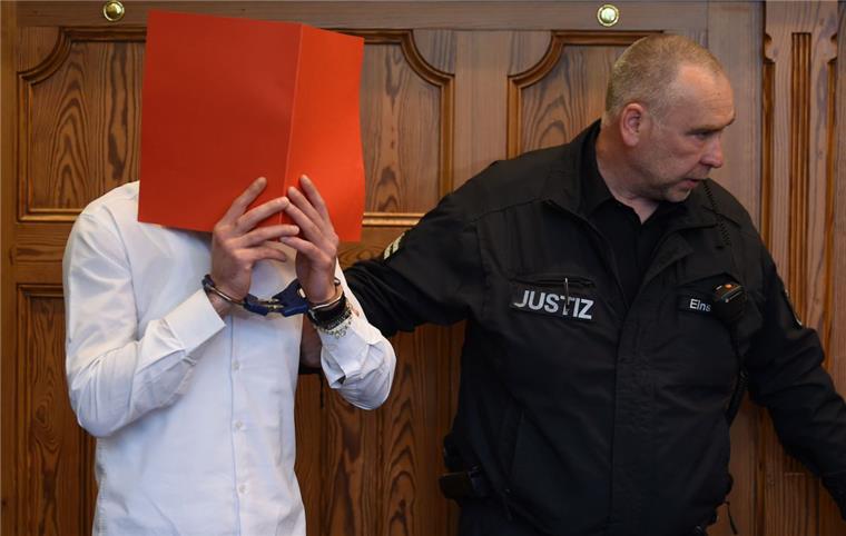 Der wegen versuchten Mordes Angeklagte wird von einem Justizbeamten im Gerichtssaal auf seinen Platz begleitet und hält sich einen Aktenordner vor das Gesicht.

