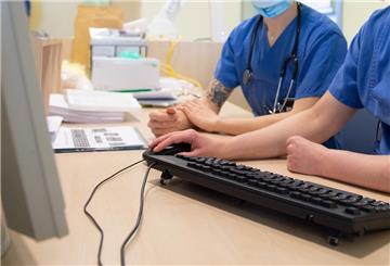 Zwei Mediziner mit blauen Kitteln und Stethoskop sitzen vor einem Computer. Die eine Person bedient die Computermaus.