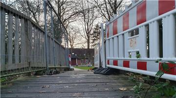 Eine Brücke führt in einen begrünten Garten. Links stehen auf der Brücke Bauzäune auf der rechten Seite stehen Baustellenbaken.