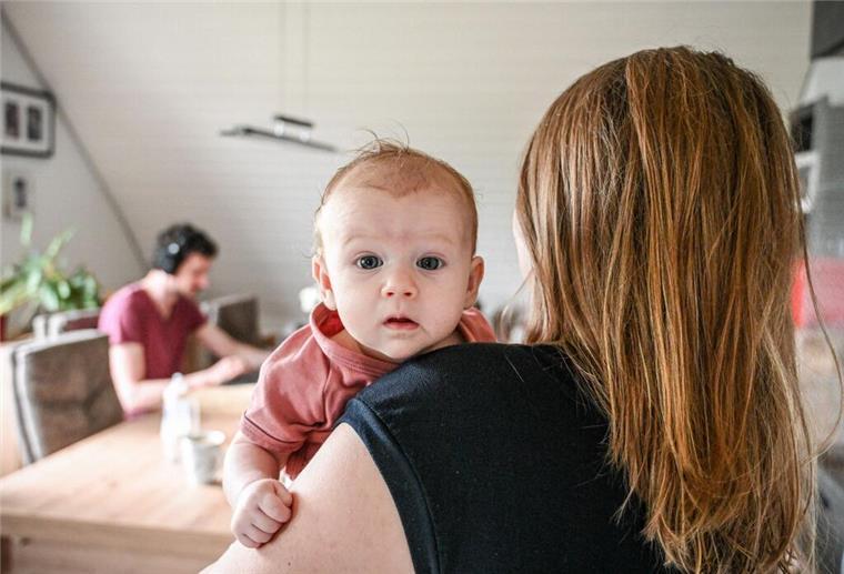 Ein Baby schaut über die Schulter der Mutter. Im Hintergrund sitzt der Vater am Esstisch der Wohnung und arbeitet am Laptop.