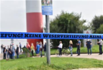Hinter einem blauen Flatterband mit der Aufschrift "Keine Weservertiefung!" sind Menschen und ein Leuchtturm zu sehen.