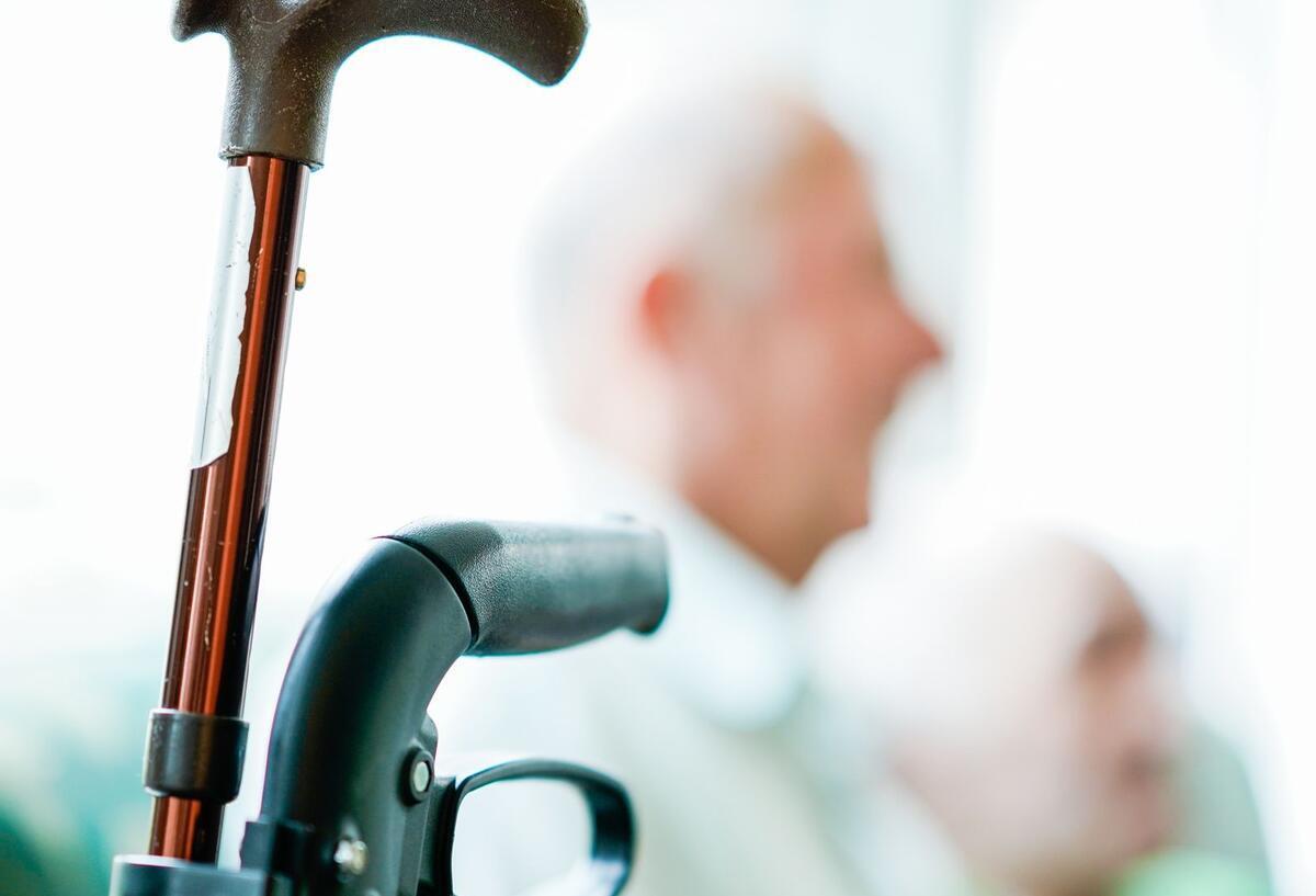Der Teil eines Rollators und eines Gehstocks. In Hintergrund sieht man unscharf einen Rentner.