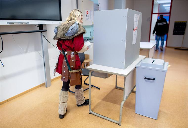 Dinosaurier, Elvis und Co. stürmen das Wahllokal! Die kostümierte Wählerin Linda geht in einem Wahllokal zur Stimmabgabe hinter einen Sichtschutz.