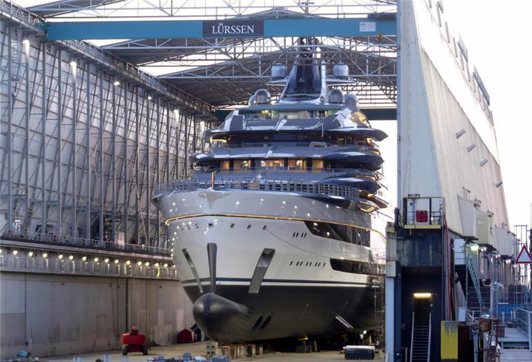 Eine riesige, luxuriöse Megayacht mit glänzendem Rumpf steht im überdachten Schwimmdock der Lürssen-Werft in Bremen-Aumund. Das Schiff ist vollständig gebaut, mehrere Decks mit umlaufenden Balkonen und Antennenaufbauten sind sichtbar. Um das Dock herum stehen technische Anlagen und Gerüste.