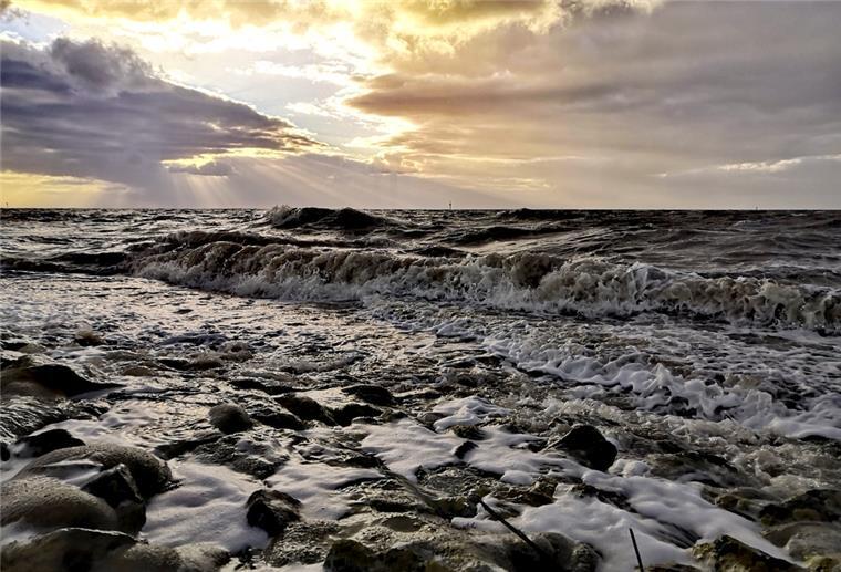 Schönes Schietwetter - nirgends scheint der Himmel so schön grau wie bei uns Die tobende Nordsee. Darüber brechen Sonnenstrahlen durch die grauen Wolken.