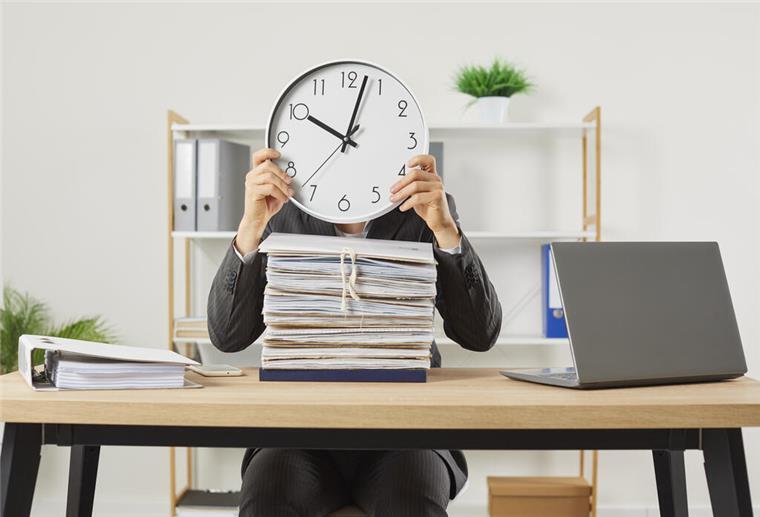 Tschüss 8-Stunden-Tag? Fast die Hälfte der Deutschen will‘s flexibler Person in Anzug sitzt an Schreibtisch mit einem großen Stapel Akten und hält eine Uhr vor das Gesicht, die zehn Minuten nach zehn anzeigt; Symbolbild für Zeitdruck oder Überlastung im Büro.