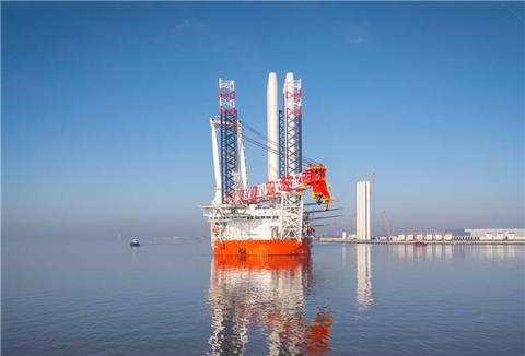 Offshore-Errichterschiff mit leuchtend orangefarbenem Rumpf und großem Kranaufsatz liegt bei klarem Himmel in ruhigem Hafenwasser; im Hintergrund sind Hafenanlagen und Windkraftkomponenten zu sehen.