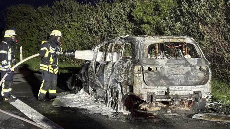 Zwei Feuerwehrleute stehen neben einem ausgebrannten Auto und besprühen es mit Löschschaum.