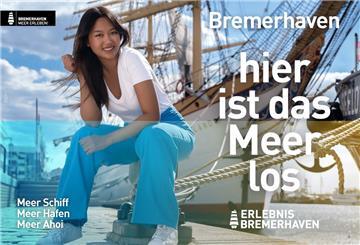 Eine junge Frau mit blauer Hose sitzt vor einem Schiff und lächelt in die Kamera.