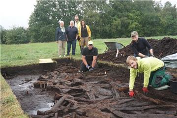 Ein Archäologenteam legt einen Streckenabschnitt des historischen Moorwegs frei. Im Hintergrund beobachten Dr. Stefan Hesse (hinten) und Dr. Marion Heumüller (rechts), Fachbereichsleiterin im Landesamt für Denkmalpflege, gemeinsam mit dem Ehepaar Specht die Arbeiten von Lennart Picht (rechts), Ingo Neumann und Jetta Harms.