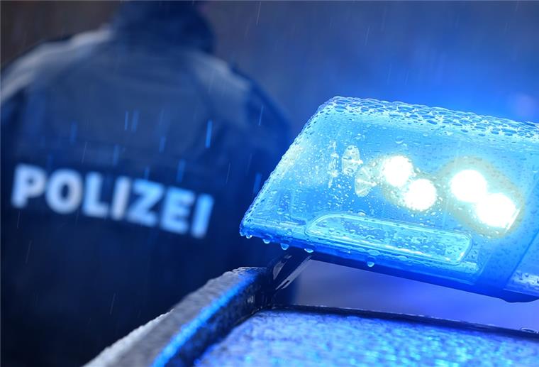 Ein Polizist steht im Regen vor einem Streifenwagen dessen Blaulicht aktiviert ist.