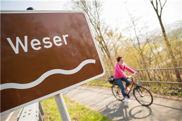 Ein Fahrradfahrer fährt auf dem Weser-Radweg vorbei an einem Wegweiser, an dem unter anderem ein Schild mit der Aufschrift "Weser".