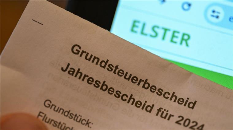 400.000 Beschwerden nach versendeten Messbescheiden Ein Grundsteuerbescheid für 2024 wird vor einen Computerbildschirm gehalten, auf dem das Logo der Steuerplattform Elster angezeigt wird.