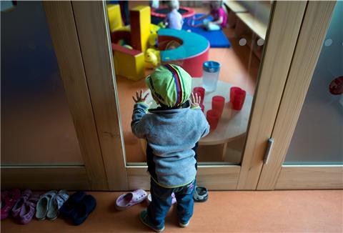 Ein Kleinkind steht an einer Glasscheibe in einer Kindertagesstätte und schaut anderen Kindern beim Spielen zu. Wegen der Pandemie laufen die Kitas nur im Notbetrieb. Normalität wünschen sich auch in Hepstedt immer mehr Eltern und Kinder. 