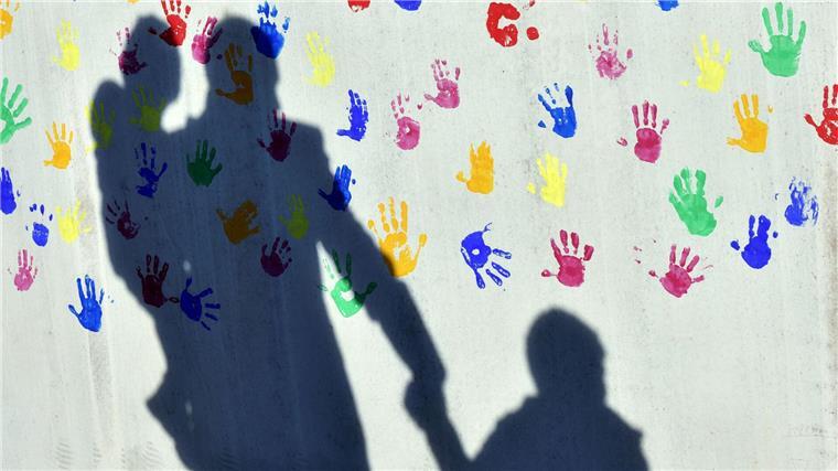Kinderschutzbund: Mehr Entschlossenheit im Kampf gegen Armut Ein Mann mit einem Kind auf dem Arm und einem an der Hand wirft einen Schatten auf eine mit bunten Handabdrücken bemalte Wand einer Kindertagesstätte. (Symbolbild)