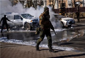 Ein ukrainischer Soldat bezieht Stellung vor einer Militäreinrichtung, während Feuerwehrleute im Hintergrund zwei brennende Autos löschen. Russische Truppen haben den erwarteten Angriff auf die Ukraine gestartet und drangen in die Hauptstadt vor. 