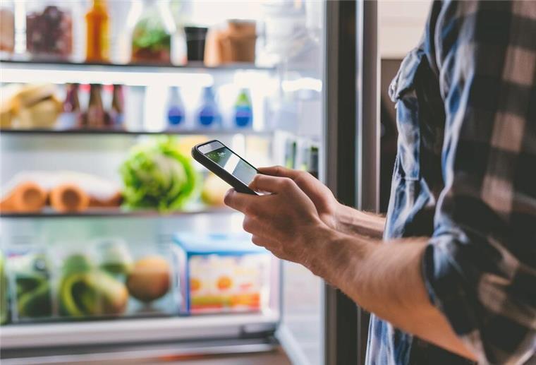 Haushalt digital organisieren: Wie smarte Helfer den Alltag erleichtern Mann steht mit Handy vor dem offenen Kühlschrank.