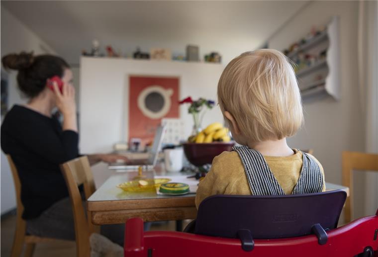 Kinderschutzbund: Wie sich die Corona-Pandemie auf Kinder auswirkt Eine Frau sitzt im Homeoffice an ihrem Laptop und telefoniert, während ihr Kind neben ihr in einem Kinderstuhl am Tisch sitzt – eine Situation, die Eltern ihren Kindern erklären sollten.