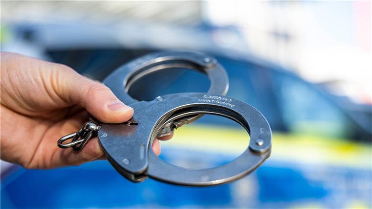 Eine Hand hält Handschellen vor einen Streifenwagen der Polizei.