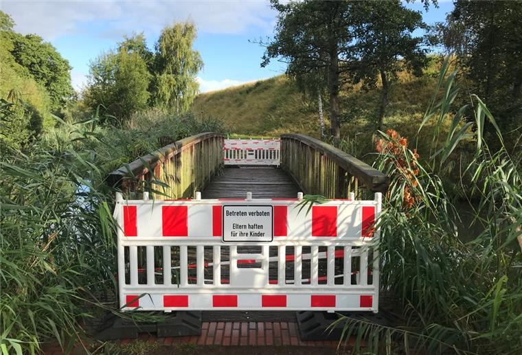 Brake: Fußgängerbrücke wird saniert Zwei rot-weiße Absperrungen verhindern den Zugang zu einer Holzbrücke, die sich inmitten grüner Natur befindet.