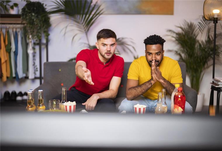 Superbowl: Was der Schlafentzug mit unserem Körper macht Zwei junge Männer sitzen vor dem Fernseher, vor ihnen stehen Snacks und Getränke auf dem Tisch.