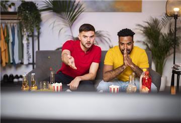 Zwei junge Männer sitzen vor dem Fernseher, vor ihnen stehen Snacks und Getränke auf dem Tisch.