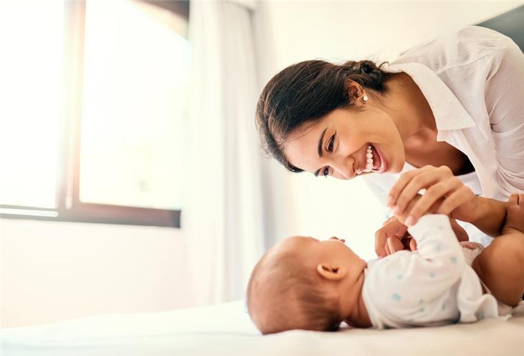 Gutartige Tumore auf zarter Babyhaut Eine dunkelhaarige Frau lacht ein auf dem Rücken liegendes Baby an.