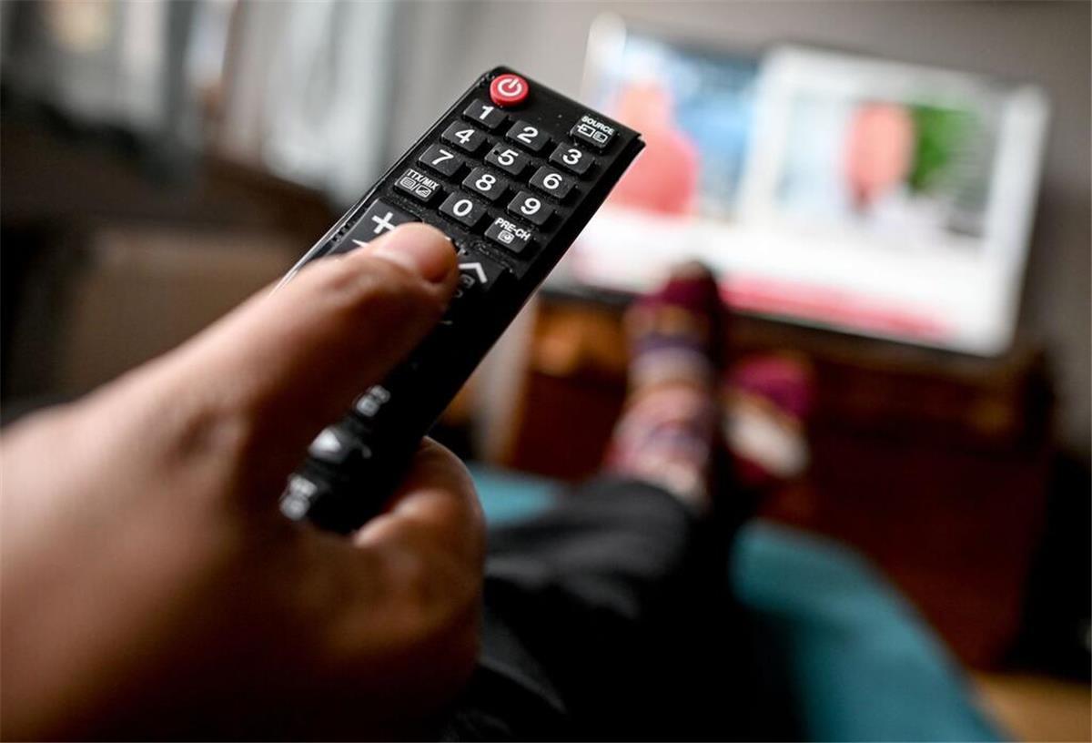 Eine Person hält eine Fernbedienung in der Hand und schaut Fernsehen. 