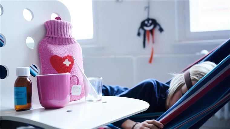 Mütter melden sich häufiger kindkrank als Väter Fieberthermometer, Hustensaft, Wärmflasche und eine Tasse Tee stehen auf einem Stuhl, im Hintergrund liegt ein krankes Kind.
