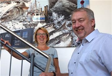 Ein Mann und eine Frau stehen auf einer Treppe. Im Hintergrund hängt ein Bild, auf dem Fische und Eis gemalt sind.
