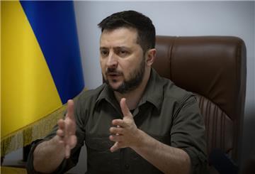 Der ukrainische Präsident Wolodymyr Selenskyj sitzt an einem Tisch und spricht. Im Hintergrund die ukrainische Flagge.